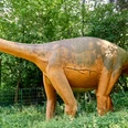 Lebensgroße Dinosaurierfigur in einem von Bäumen umgebenen Grünstreifen.