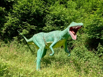 Grüne Dinosaurier-Skulptur mit offenem Maul steht in lichter Waldumgebung.Green dinosaur sculpture with open mouth stands in a light forest setting.Grøn dinosaurskulptur med åben mund står i lyse skovomgivelser.Groen dinosaurusbeeld met open bek staat in een lichte bosrijke omgeving.