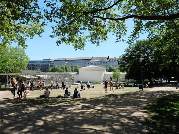 Mehrere Personen spazieren oder sitzen auf der Wiese in einem Park mit Blick von hinten auf das Gebäude des Elisenbrunnens.