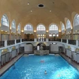 Historisches Hallenbad mit hohem Gewölbe, Arkadenfenstern und zentralem Schwimmbecken.