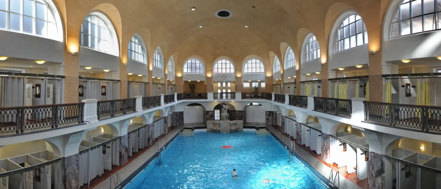 Historisches Hallenbad mit hohem Gewölbe, Arkadenfenstern und zentralem Schwimmbecken.