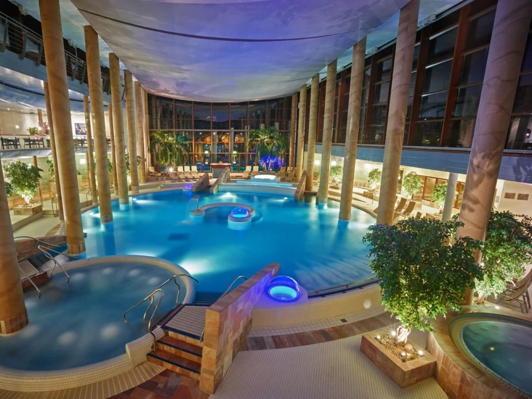 Carolus Thermen Großes, rundliches Thermenbecken umgeben von Säulen und Liegen. Beleuchtung und Pflanzen schaffen eine entspannte Atmosphäre.