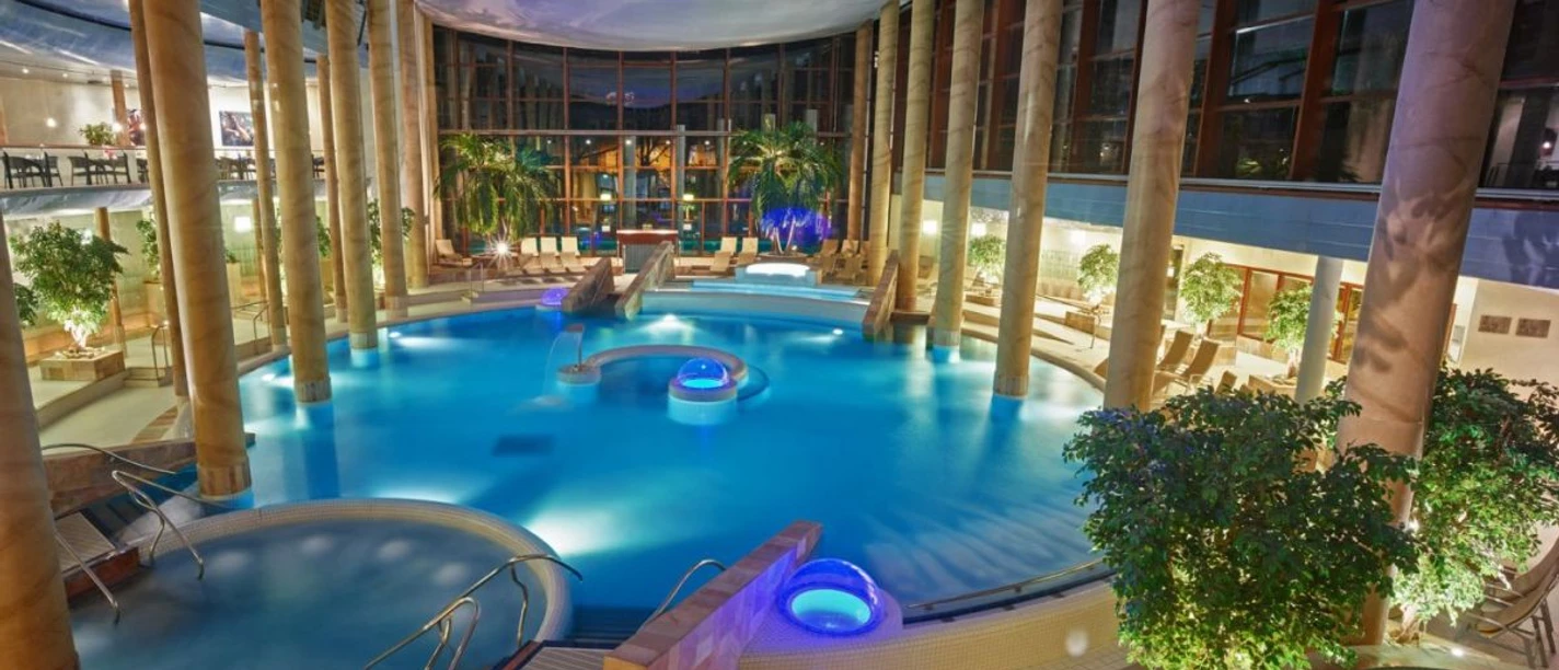 Carolus Thermen Großes, rundliches Thermenbecken umgeben von Säulen und Liegen. Beleuchtung und Pflanzen schaffen eine entspannte Atmosphäre.