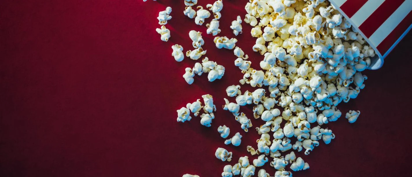 Ein umgefallener Popcornbecher mit rot-weißen Streifen, Popcorn verteilt sich auf rotem Hintergrund.