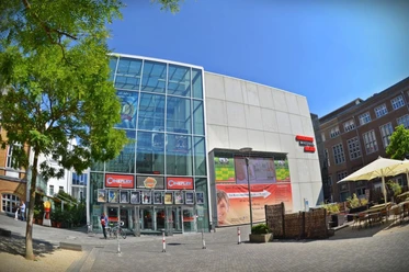 Glasfront des Kino-Gebäudes mit Werbeplakaten und einem sonnigen Platz davor.