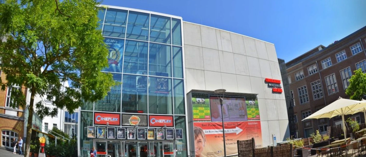 Cineplex Glasfront des Kino-Gebäudes mit Werbeplakaten und einem sonnigen Platz davor.