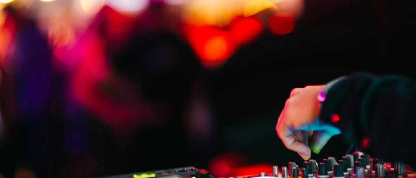Eine Hand dreht an einem DJ-Mischpult, umgeben von farbenfrohen Lichtern und verschwommenen Silhouetten auf einem Dancefloor.