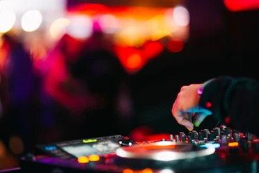 NOX Eine Hand dreht an einem DJ-Mischpult, umgeben von farbenfrohen Lichtern und verschwommenen Silhouetten auf einem Dancefloor.