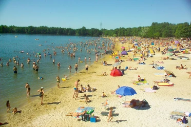 Dürener Badesee Strand Sandstrand mit vielen Besuchenden, die teils im See sind und teils am Ufer mit Sonnenschirmen liegen.