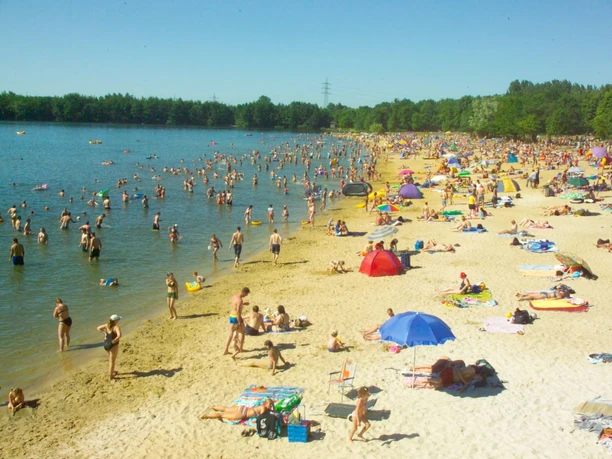 Dürener Badesee Strand Sandstrand mit vielen Besuchenden, die teils im See sind und teils am Ufer mit Sonnenschirmen liegen.