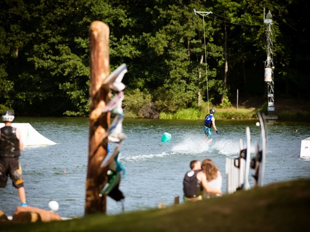 Dürener Badesee Wasserski Seitlich stehen Menschen mit Schwimmwesten und Helmen, die einen Wassersportler beobachten.
