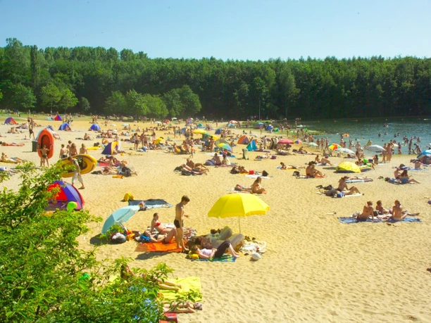 Viele Menschen liegen mit Sonnenschirmen auf dem sandigen Ufer, einige sind im Wasser schwimmen.