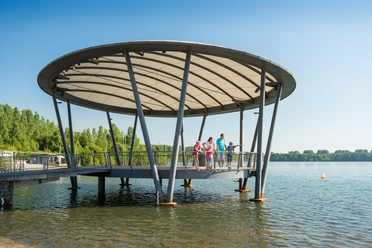 Eine Familie genießt den sonnigen Tag auf einem modernen Steg über dem Blausteinsee.