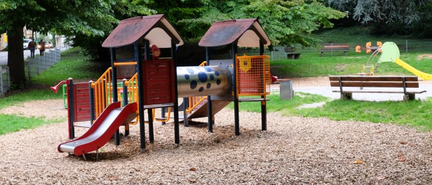 Spielplatz Veltmanplatz (c) ats Spielplatz auf Holzspänen mit einem bunten Klettergerüst, einer Rutsche und mehreren Holzbänken.