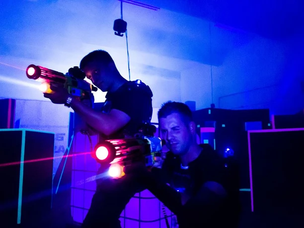Zwei Personen hocken mit LaserTag-Gewehren in einem blau belichtetem Raum vor Hindernissen.