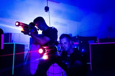 Black LaserTag Aachen Zwei Personen hocken mit LaserTag-Gewehren in einem blau belichtetem Raum vor Hindernissen.