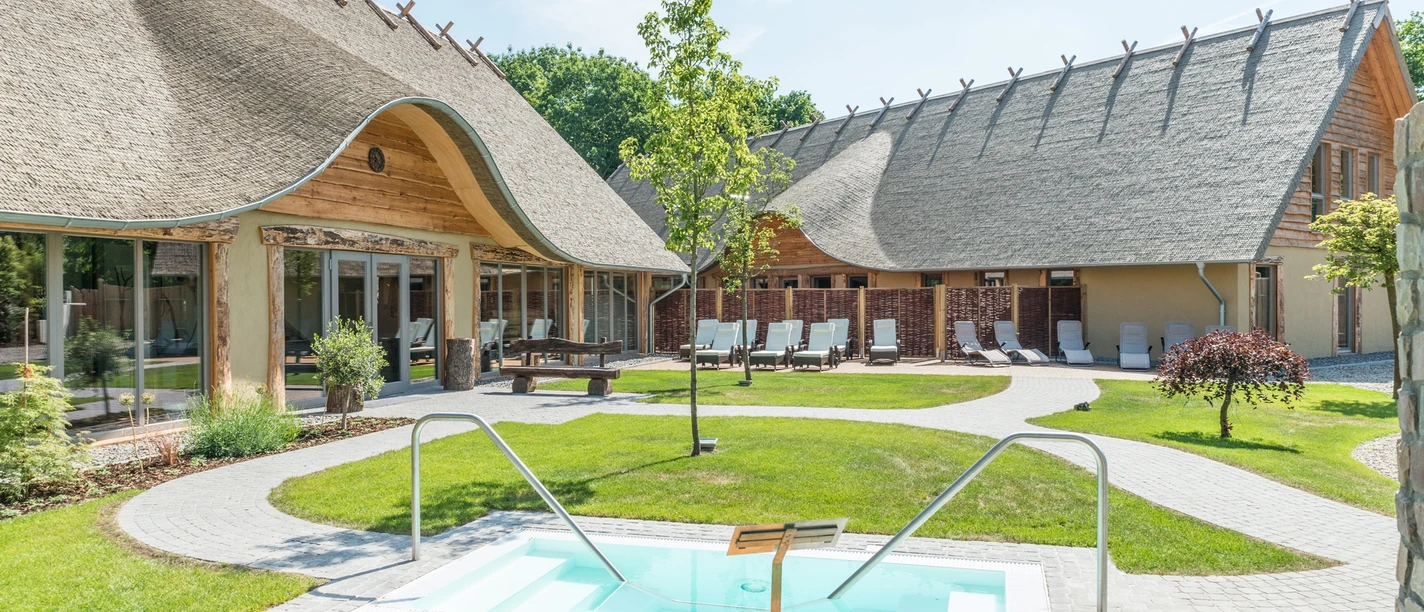 Im Stil der Germanen: Das Alfen Saunaland Outdoor area of a rural wellness hotel with thatched roof, pool and sun loungers.