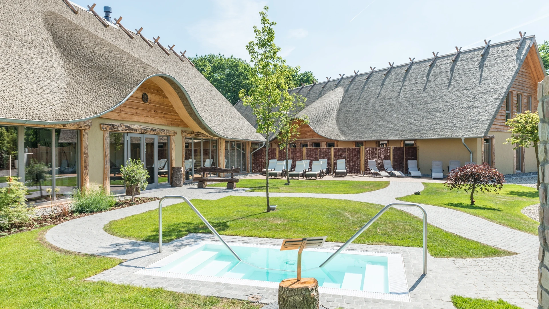 Außenbereich eines ländlichen Wellnesshotels mit Reetdach, Pool und Liegestühlen.