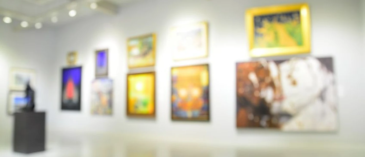 In einem modernen Galerieraum hängen verschwommene, farbenfrohe Kunstwerke an einer weißen Wand, darunter ein impressionistisches Bild eines Hundes. Die Decke ist mit Lichtstrahlern ausgestattet, die den Raum hell erleuchten.