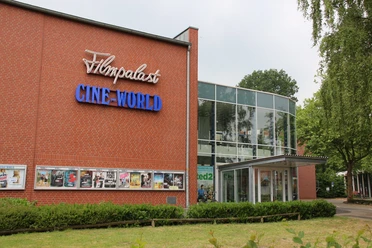 Backsteinbau mit Glasfassade und Leuchtschrift Filmpalast Cine-World, umgeben von Bäumen und Grünflächen.