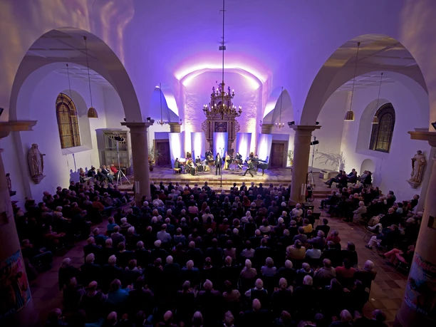 Ehemalige Kirche Hagen a.T.W._musica viva_CC_Herbert Vieth Konzert in einer Kirche mit violett beleuchteten Bögen und voll besetzten Zuschauerrängen.