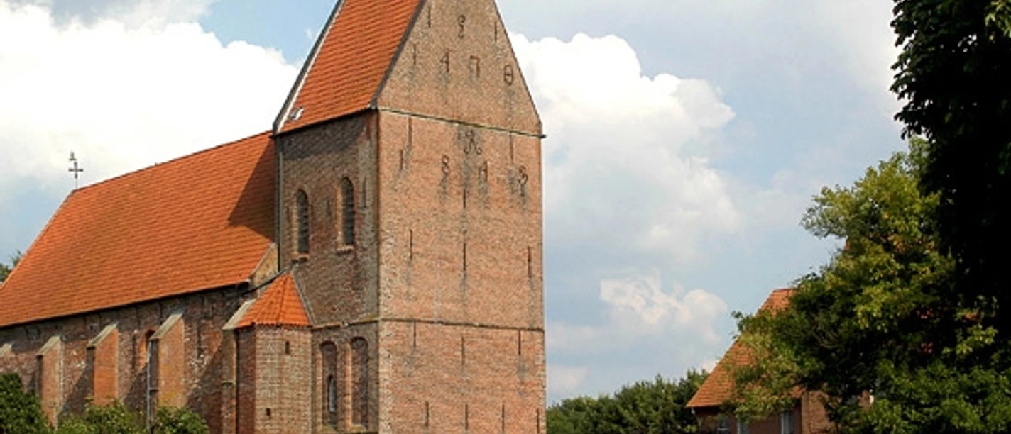 Gedächniskirche Rhede Backsteinkirche mit hohem Turm und rotem Ziegeldach, umgeben von Bäumen und blauem Himmel.