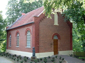 Alte Schule Bokeloh Backsteingebäude mit Satteldach, hohen Rundbogenfenstern und Statue vor dem Eingang, umgeben von Bäumen.