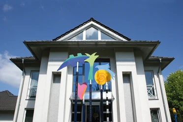 AtelierSpuren Meppen Modernes Gebäude mit farbenfroher Metallskulptur aus abstrakten Formen vor blauem Himmel.