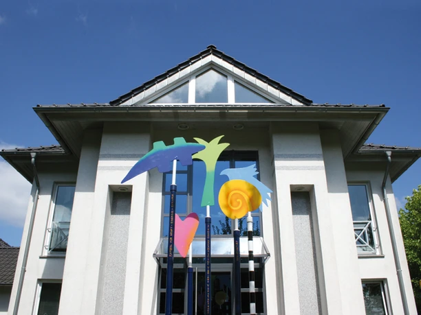AtelierSpuren Meppen Modernes Gebäude mit farbenfroher Metallskulptur aus abstrakten Formen vor blauem Himmel.