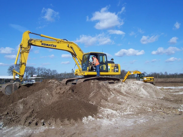 Bagger Park Haren Gelber Bagger auf sandigem Gelände mit Mensch in Sicherheitsweste unter blauem Himmel.