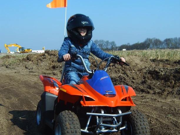 Kinderquad fahren Kind fährt mit Helm auf rotem Quad über eine sandige Piste unter blauem Himmel.