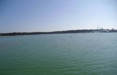 Baggersee Walchum Weitläufiger Baggersee mit ruhigem Wasser und Sandhügeln im Hintergrund unter klarem Himmel.