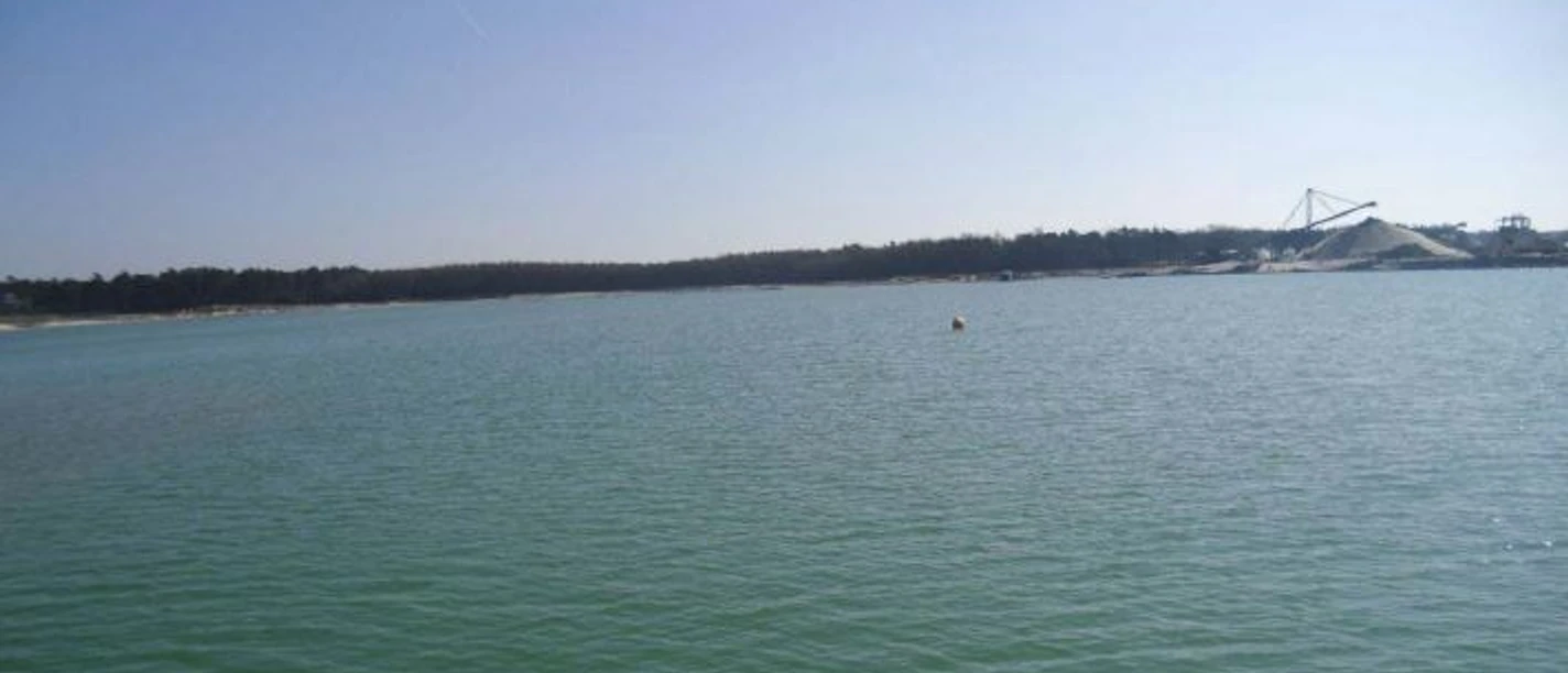 Baggersee Walchum Weitläufiger Baggersee mit ruhigem Wasser und Sandhügeln im Hintergrund unter klarem Himmel.