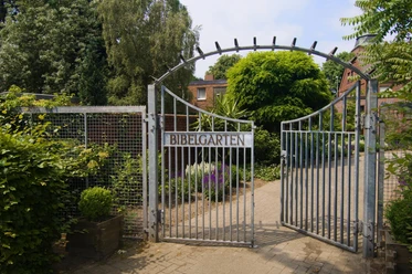 Bibelgarten Werlte Metalltor mit der Aufschrift „Bibelgarten“, dahinter blühende Beete und dichte, grüne Bäume.