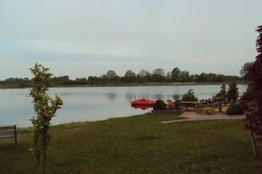 Blaue Lagune Ruheraum am Seeufer mit rotem Tretboot, grüner Wiese und Sitzbänken vor ruhigem Wasser.