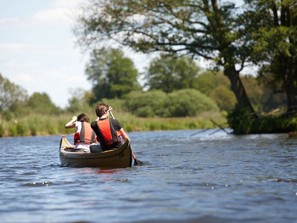 Kanufahren_Ems_Hase_02.jpg Zwei Personen paddeln in einem Kanu über einen ruhigen Fluss, umgeben von grüner Landschaft.