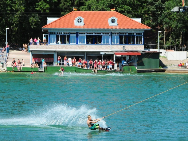 d30_3951 Wakeboardfahrer gleitet über den Dankernsee, Zuschauer genießen Sonne am Ufer vor einem Gebäude.