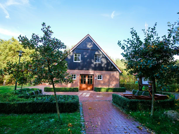 dorfscheune-varloh-au-enansicht-02-emsland-tourismus-gmbh-birgit-janknecht Backsteinhaus mit dunklem Giebel, rotem Ziegeldach und gepflegtem Vorplatz im Grünen.