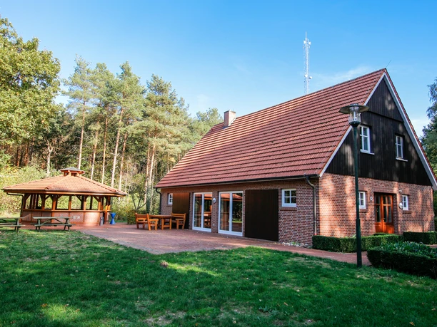 dorfscheune-varloh-au-enansicht-03-emsland-tourismus-gmbh-birgit-janknecht Backsteingebäude mit rotem Ziegeldach und Holzpavillon vor einem Wald unter blauem Himmel.