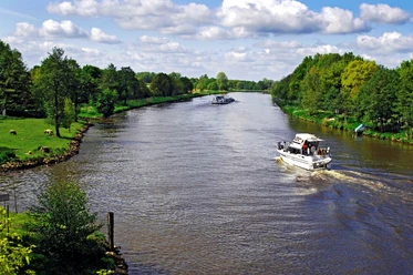 Motorboot fährt auf der Ems zwischen grünen Uferwiesen und dichten Bäumen unter blauem Himmel.