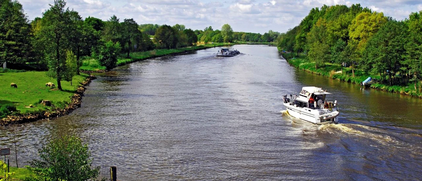 Motorboot fährt auf der Ems zwischen grünen Uferwiesen und dichten Bäumen unter blauem Himmel.