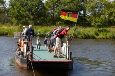 Emsfähre Leher Pünte Personen mit Fahrrädern auf kleiner Fähre über die Ems, im Wind weht eine Deutschlandflagge.