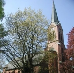 Rote Backsteinkirche mit hohem Turm und spitzem Dach, umgeben von Bäumen unter blauem Himmel.