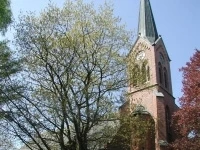 Rote Backsteinkirche mit hohem Turm und spitzem Dach, umgeben von Bäumen unter blauem Himmel.