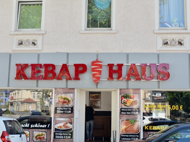 Döner-Restaurant mit auffälligem roten Schild und Glasschiebetür, Werbung für Speisen im Fenster.