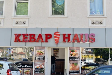 Kebap-Haus Außenansicht Döner-Restaurant mit auffälligem roten Schild und Glasschiebetür, Werbung für Speisen im Fenster.