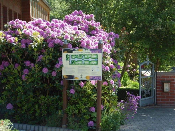Garten des Nazareners (Bibelgarten) Eingang zum Bibelgarten mit blühenden Rhododendren, Informationstafel und offenem Eisengittertor.