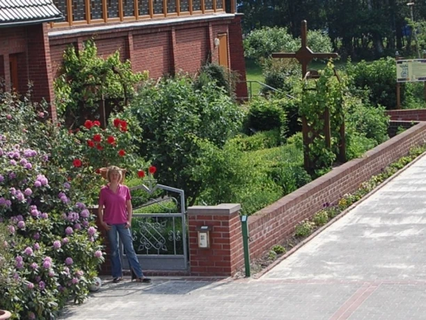 Garten des Nazareners (Bibelgarten) Frau am Eingang des Bibelgartens vor blühenden Rosen und Ziegelgebäude mit großem Holzkreuz.