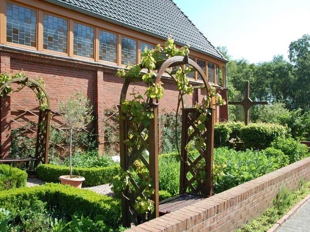 Garten des Nazareners (Bibelgarten) Grüner Bibelgarten mit Pflanzenbeeten, Holzbögen und Kreuz an roter Kirchenmauer im Sonnenlicht