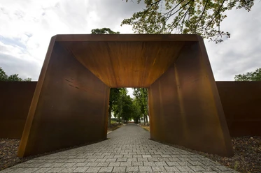 Gedenkstätte Esterwegen Rostfarbener Stahlbogen mit gepflastertem Weg, der in eine von Bäumen gesäumte Allee führt.